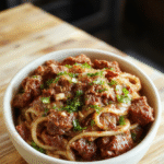 Vibrant bowl of Mongolian ground beef noodles featuring glossy, caramelized ground beef on top of tender stir-fried noodles, garnished with sliced green onions and sesame seeds, colorful sauce coating the dish, styled simply on a white plate with a neutral background.