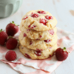 Golden-brown cookies with visible strawberry specks, dusted with powdered sugar, arranged on a white plate with fresh berries and a crumb topping, soft pastel backdrop, rustic yet inviting.