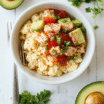 A vibrant bowl arranged on a white plate: cauliflower rice base topped with flaked tuna in a glossy spicy dressing, cucumber, cherry tomatoes, avocado slices, and green onion with sesame seeds and cilantro, all bright under natural light.