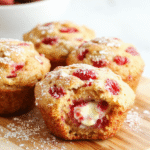 Plump moist muffins with pink strawberry specks and creamy centers, golden tops, arranged on a white plate with a light wooden background and a touch of powdered sugar